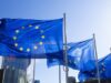 Drei EU-Flaggen mit Fahnenmast rechts wehen bei blauem Himmel in Brüssel.