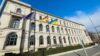 Gebäude des Bundesministeriums für Digitales und Verkehr bei Sonnenschein - vor dem Ministerium wehen die EU-, die deutsche und die ukrainische Flagge (v. l. n. r.). Auf dem Gehweg ist eine Person unterwegs.